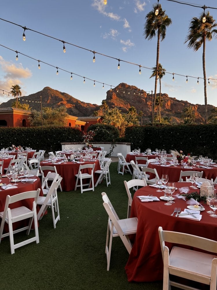Outdoor wedding reception with burgundy tablecloths and white chairs on grass, string lights overhead, and desert mountains in the background