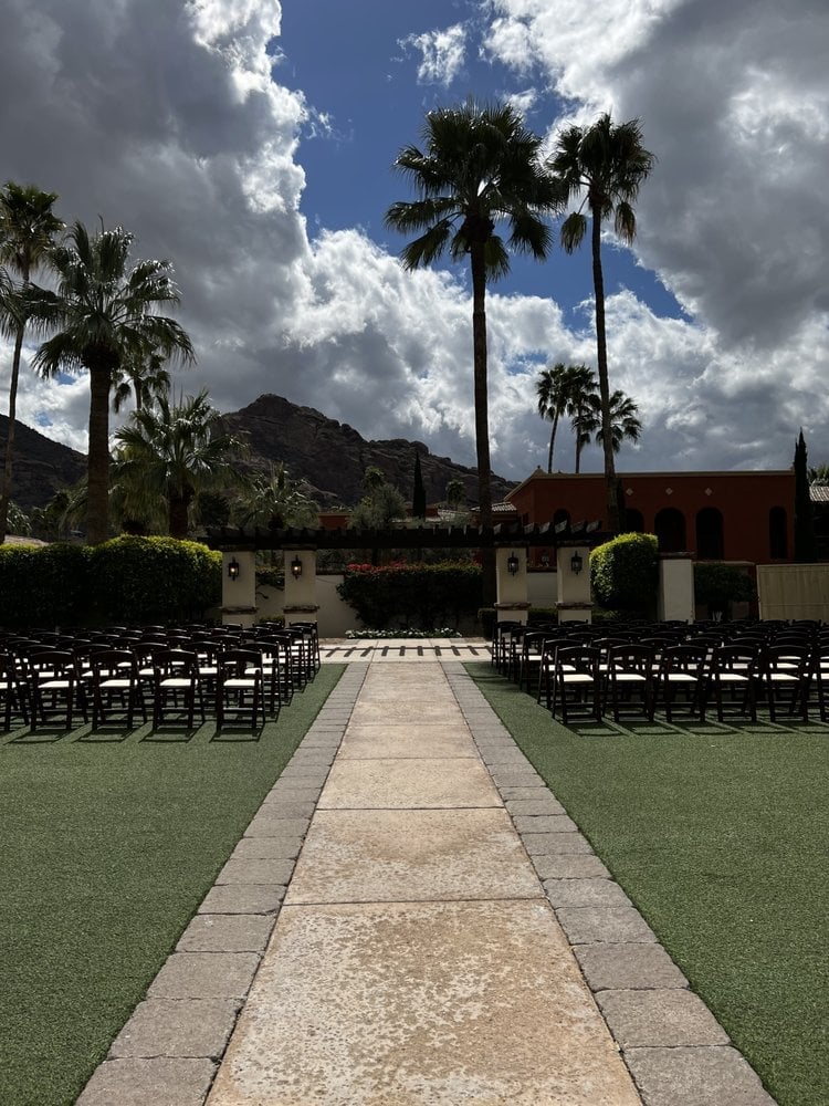 Outdoor wedding ceremony setup with stone aisle, black chairs on both sides, tall palm trees, mountains, and dramatic sky with clouds