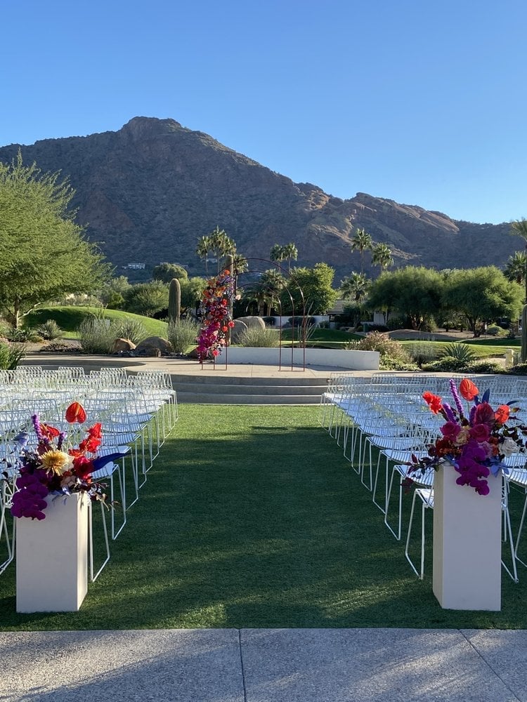 Outdoor wedding ceremony setup with white chairs lining a grass aisle, floral arrangements, and desert mountains in the background