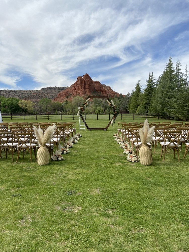Outdoor wedding ceremony setup on green lawn with arch, wooden chairs lined in rows, red rock formation and forest backdrop under blue sky