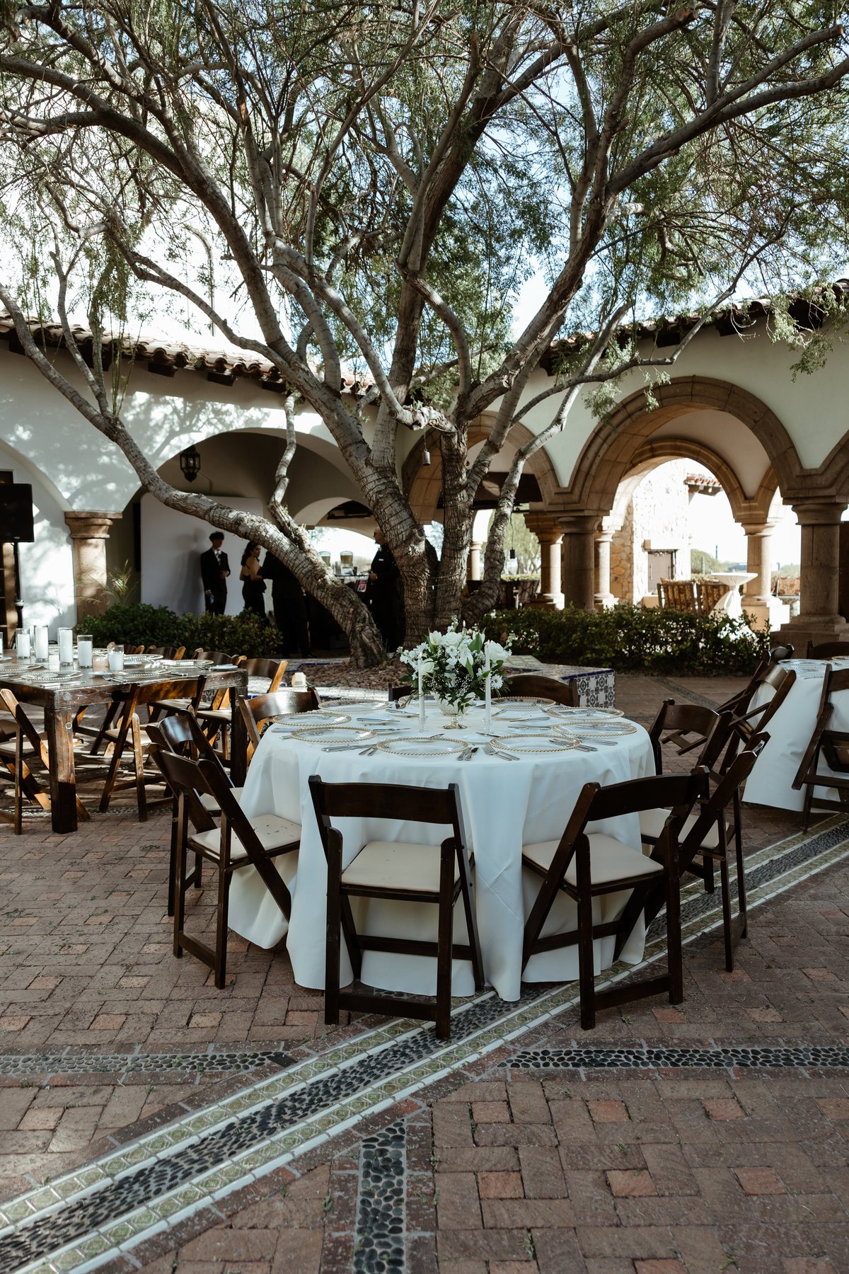 Outdoor celebration with round tables set beneath a large mature tree with Spanish colonial arches
