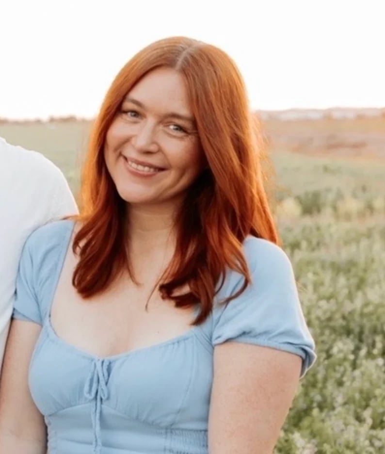 Woman with long red hair wearing a light blue top, smiling outdoors in a rural field setting
