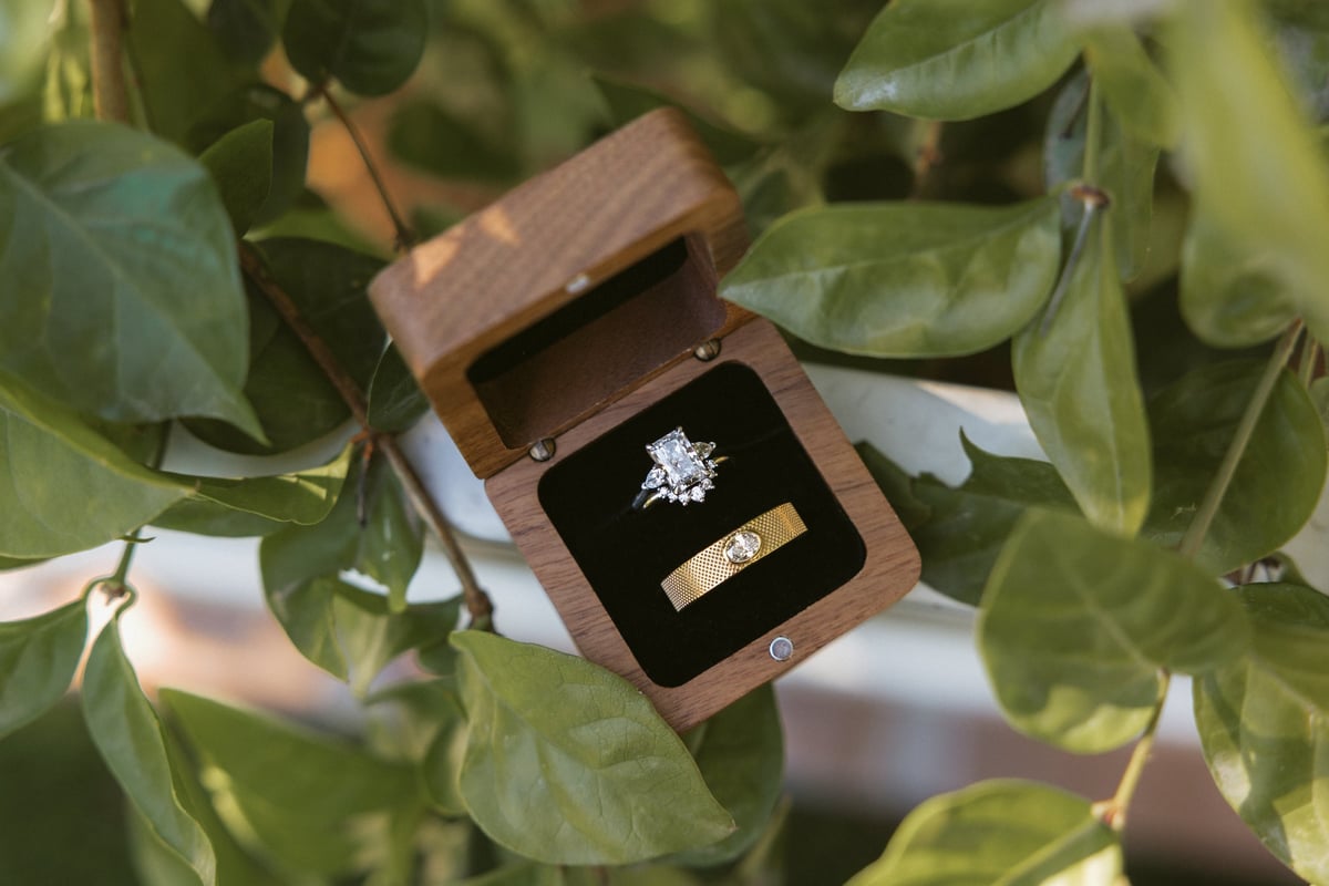 Wooden ring box with diamond engagement ring and gold wedding band among green foliage