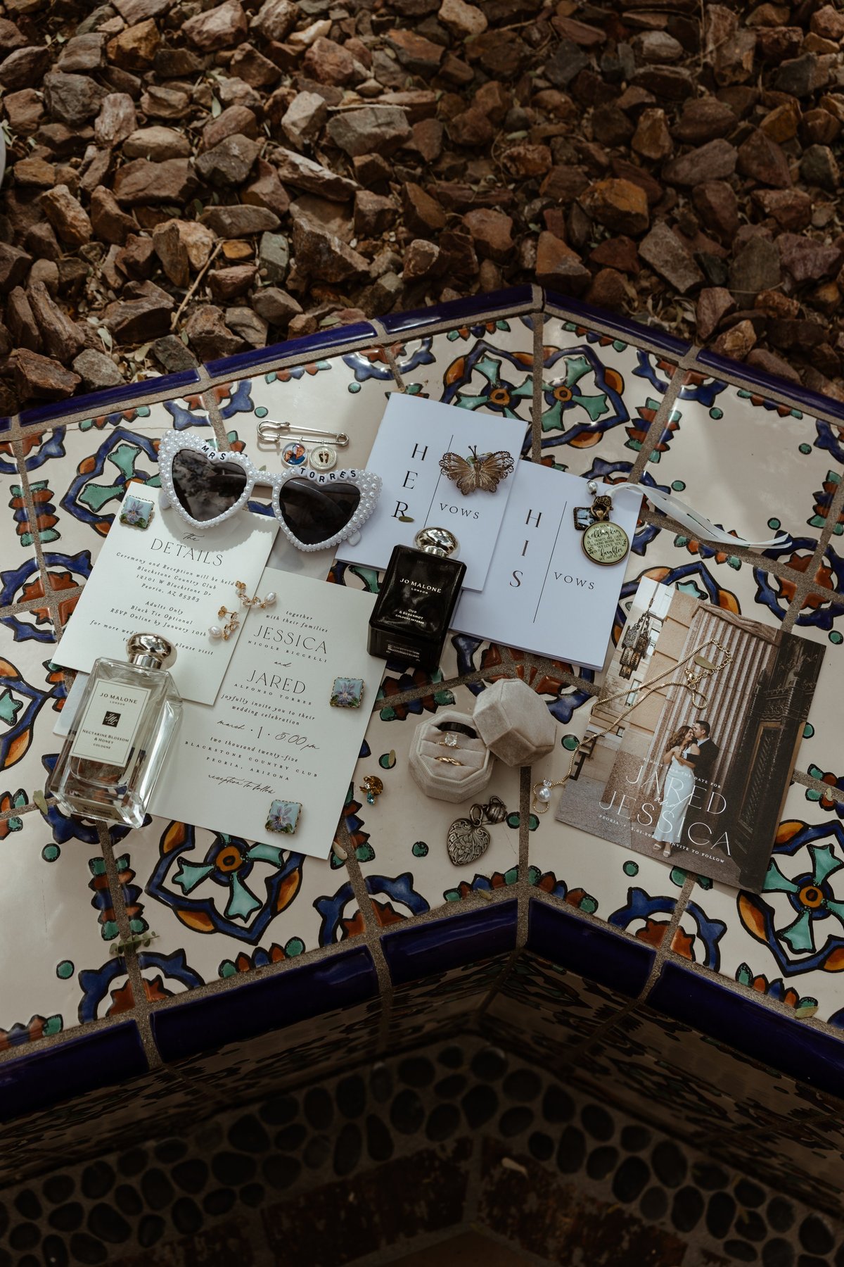Styled flat lay of wedding stationery, perfume bottles, and decorative items arranged on a patterned decorative tray with stones in background
