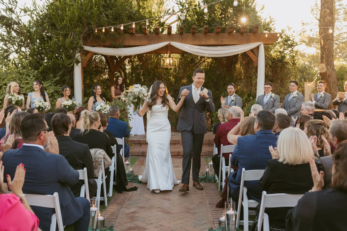 Bride and groom exchange vows at outdoor wedding ceremony under draped wooden arch with guests seated on both sides