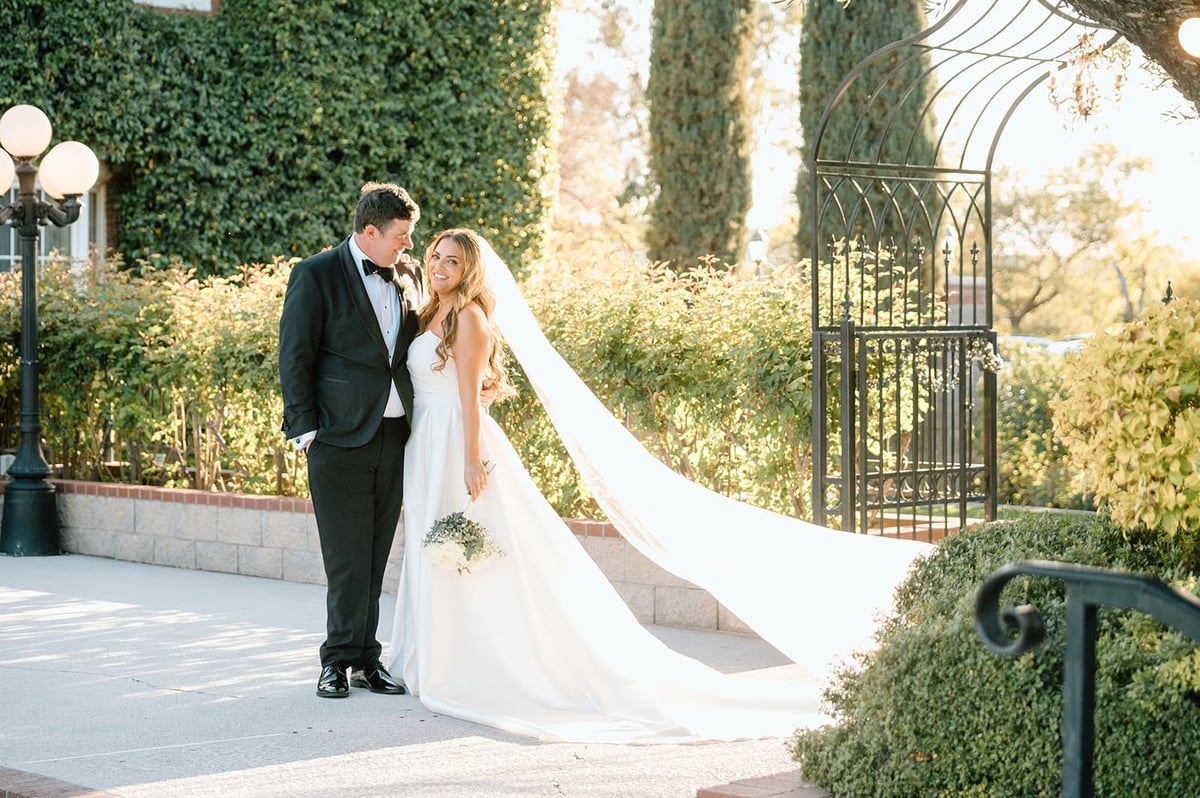 Bride and groom embrace on a stone patio with lush greenery and an ornate gate in the background
