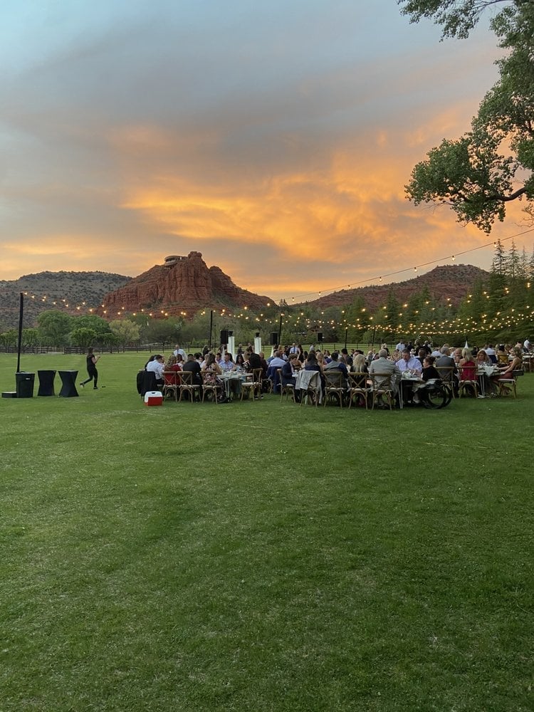 Outdoor dining event at sunset with guests seated at tables on a grassy field, string lights overhead, and desert mountains in the background