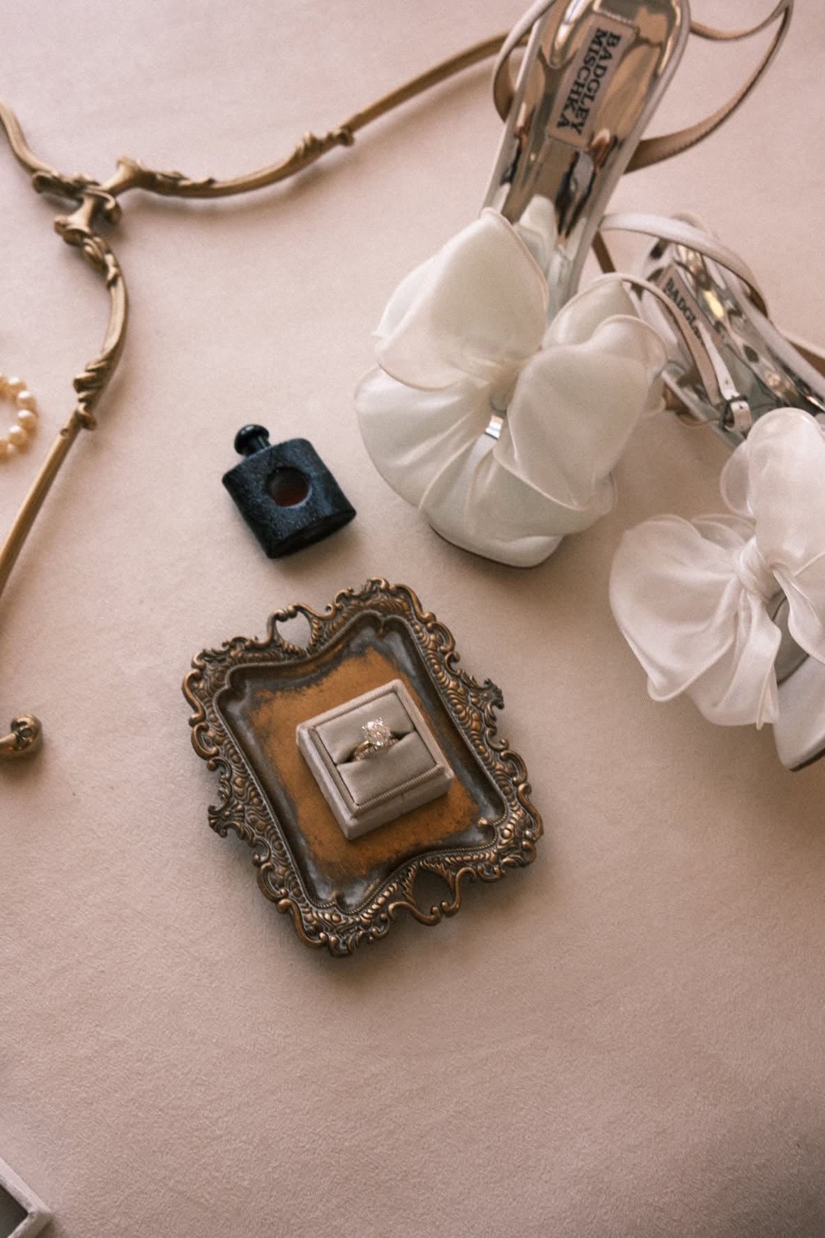 Wedding details arranged on a beige surface: ornate frame holding engagement ring, white hair clip, small blue perfume bottle, and jewelry pieces