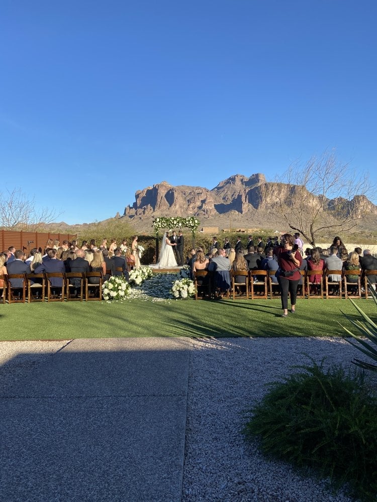 Outdoor wedding ceremony in desert setting with guests seated, dramatic mountain backdrop, white flower arrangements, and clear blue sky