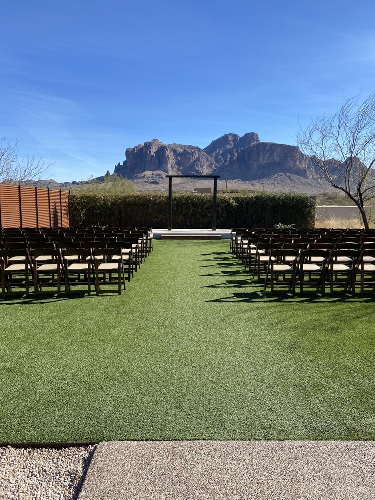 Outdoor wedding ceremony setup with black chairs on green lawn, rustic arch, and desert mountain backdrop