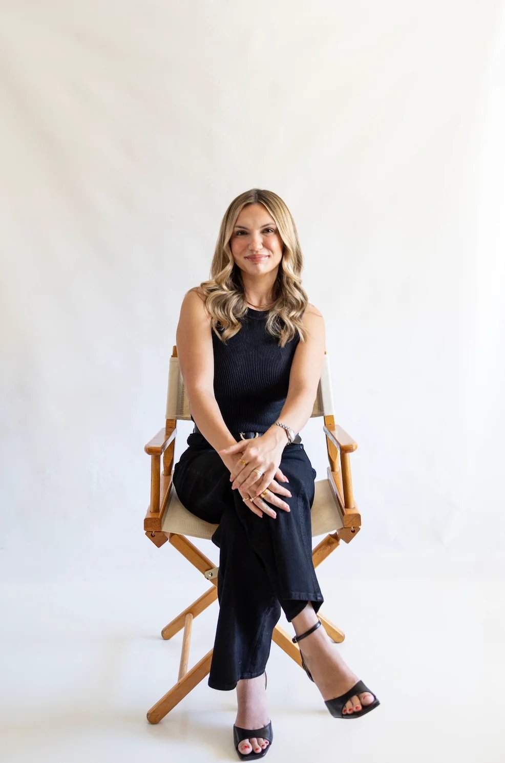 Woman with blonde wavy hair sitting on a wooden directors chair wearing a black sleeveless top and pants