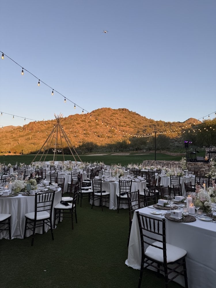 Outdoor wedding reception setup with white tablecloths, black chairs, and string lights on a green lawn with desert hills backdrop