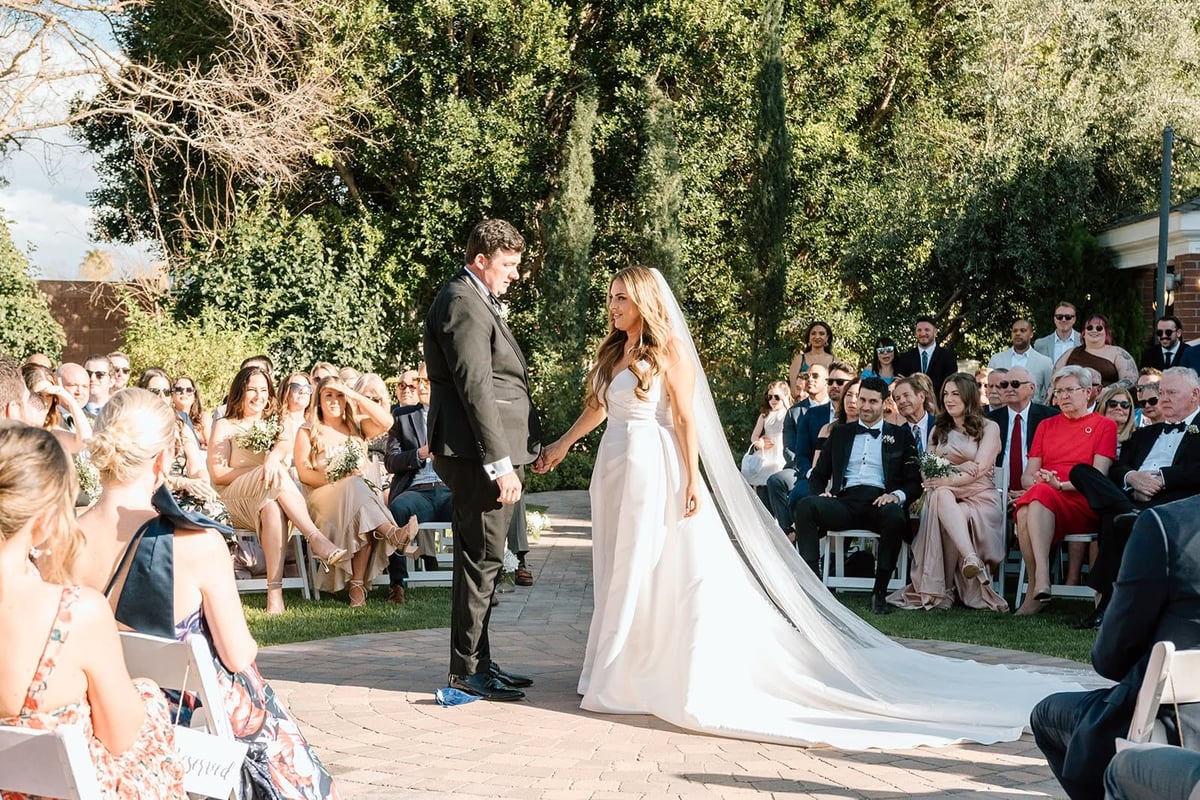 Bride and groom exchanging vows during outdoor wedding ceremony with guests seated around them on a sunny day